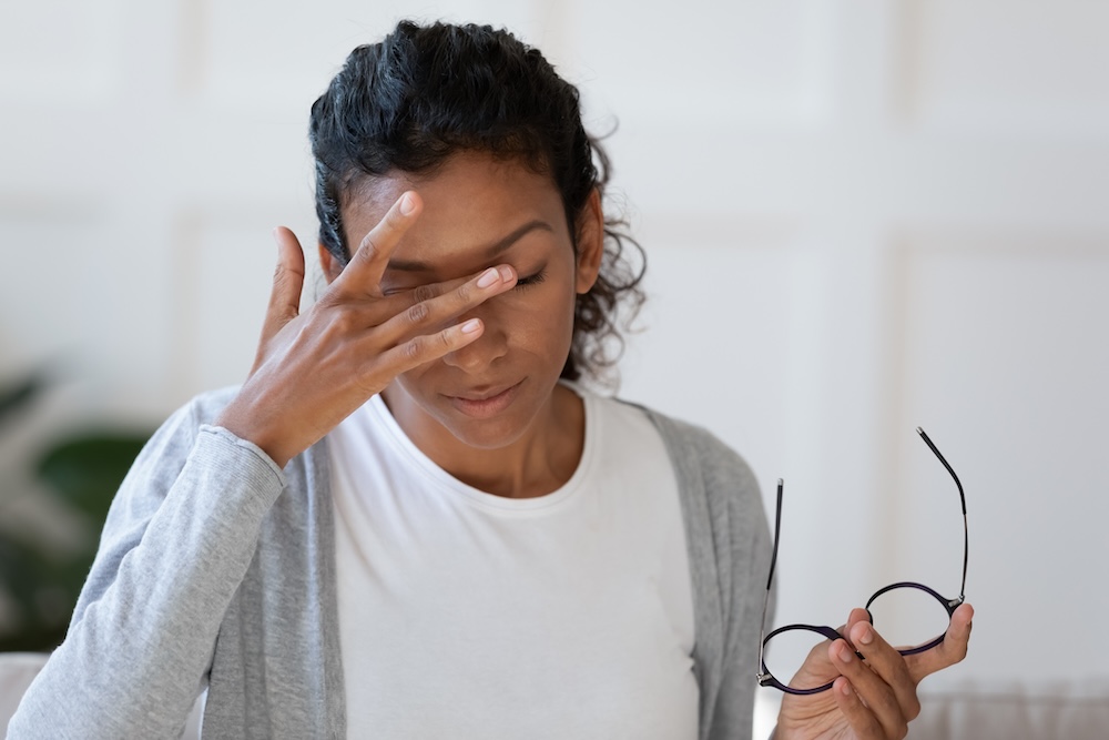 A young woman with brown skin and curly dark hair rubs her eyes due to dry eye disease symptoms while holding her glasses in the other.