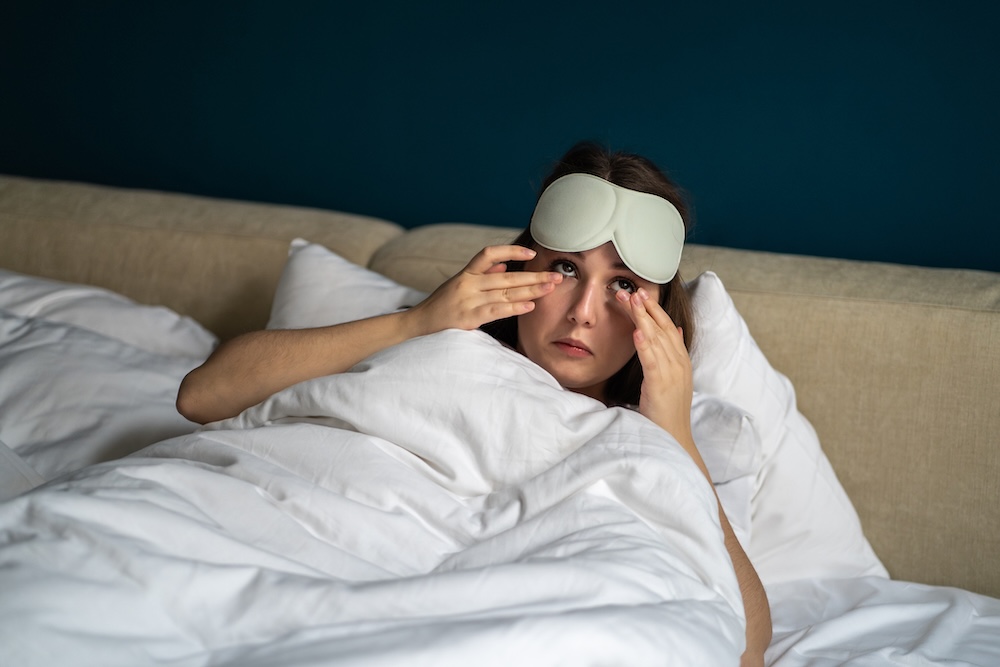 An image of a young woman in bed with an eye mask waking up with dry eyes in the morning.